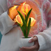 A hand holds a glowing orange LED tulip night light with a green plant in a white ceramic pot.