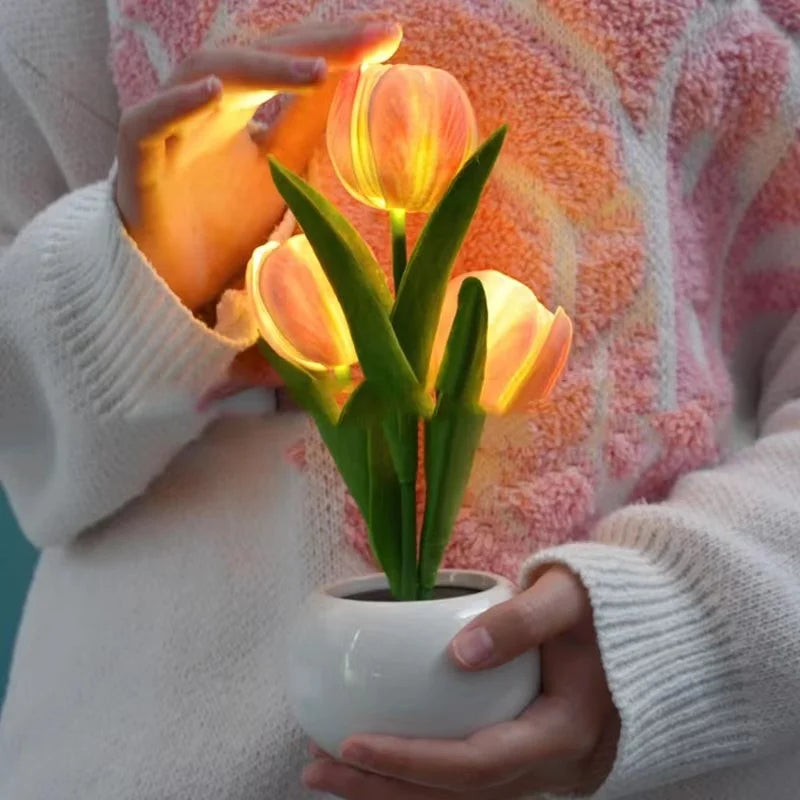 A hand holds a glowing orange LED tulip night light with a green plant in a white ceramic pot.