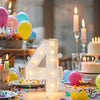 A glowing white LED marquee number 4 is displayed on a table decorated with colorful confetti and balloons for a celebration.