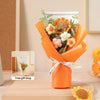 An orange gift bag tied with a white ribbon holds a bouquet of handcrafted crochet sunflowers and other flowers.