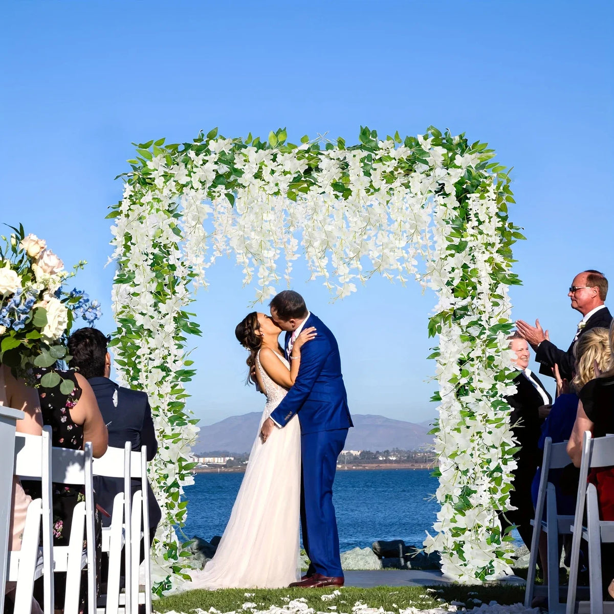 A bride and groom kiss under a white floral vine archway overlooking the sea.