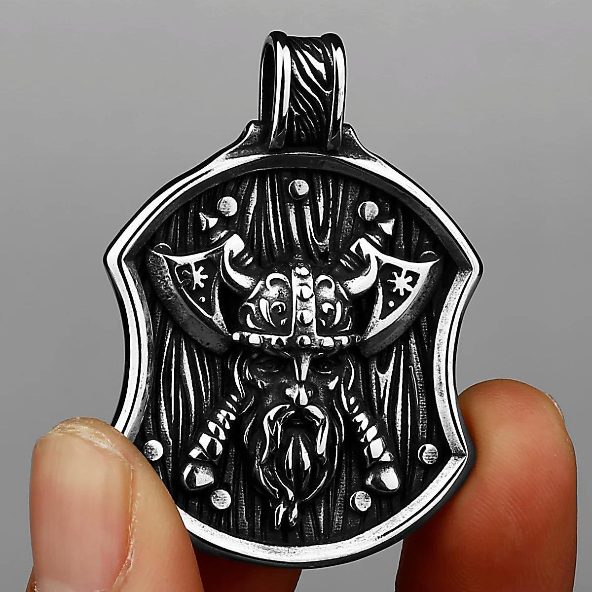 A silver stainless steel Viking warrior shield pendant is held by a hand against a gray background.