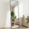 A full length floor mirror with a black frame stands in a bright room, reflecting a window and interior decor.