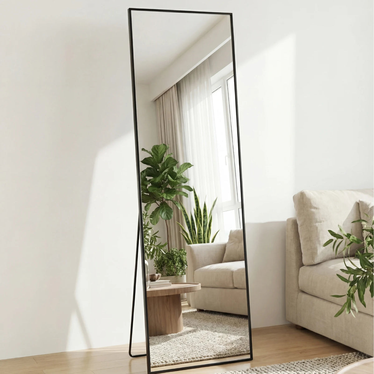 A full length floor mirror with a black frame stands in a bright room, reflecting a window and interior decor.