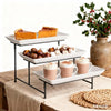 A black metal three-tier serving rack displaying baked goods and beverages on a wooden surface.