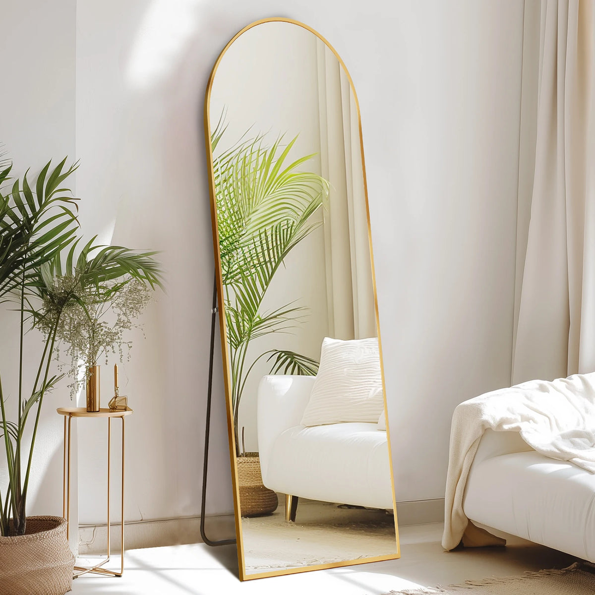 A full length arched wall mirror with a gold frame reflects a green palm leaf and interior decor.