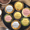 A black platter holds ten cupcakes with gold and pink birthday message toppers.