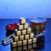 A stack of reusable stainless steel ice cubes on a dark surface next to a glass of amber liquid.