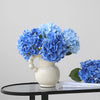A bouquet of bright blue artificial hydrangea flowers in a white vase on a black table.