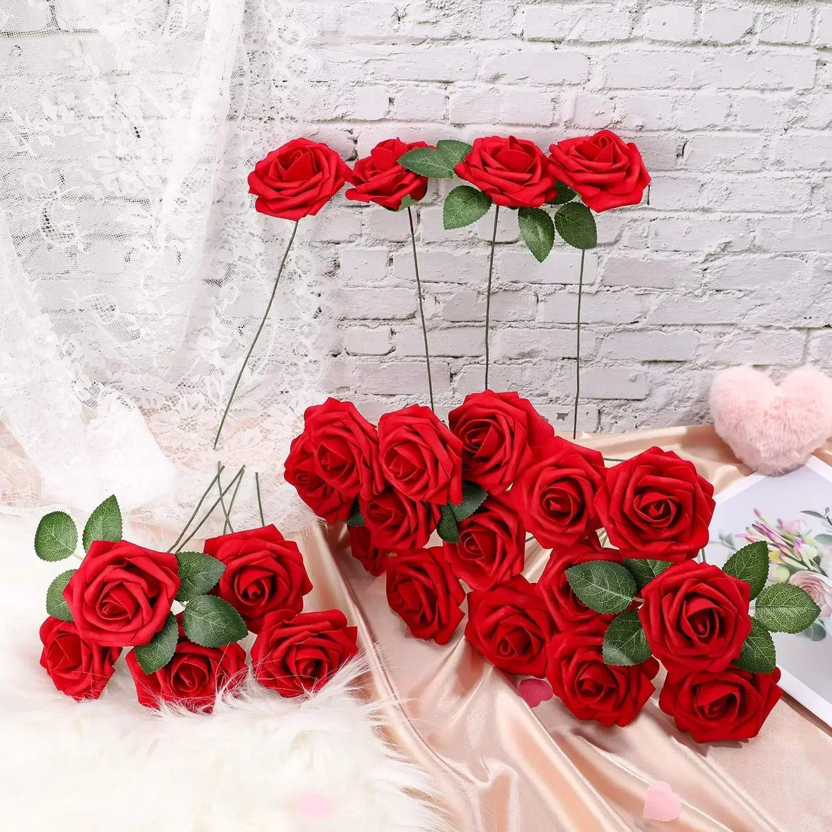 A cluster of lifelike red foam roses with green leaves displayed on a satin fabric against a white brick background.