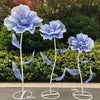 A set of three large blue velvet mesh flowers with attached leaves displayed against a green hedge.