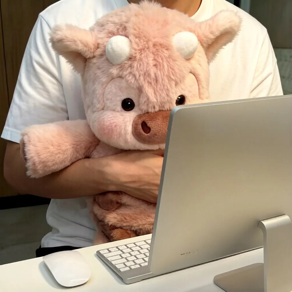 Une personne tient un jouet en peluche Highland Calf rose Kawaii et utilise un ordinateur portable argenté.