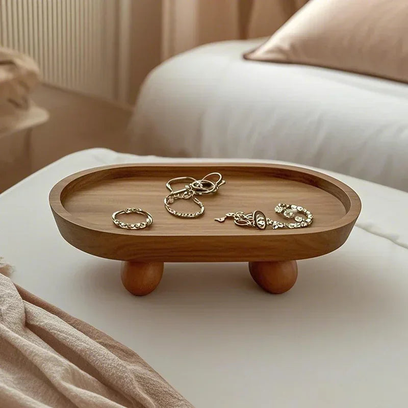 A wooden elliptical tray holds several silver-toned jewelry pieces on a white bed linen.