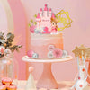 A pink tiered cake decorated with a castle topper, surrounded by floral accents and gold geometric shapes.