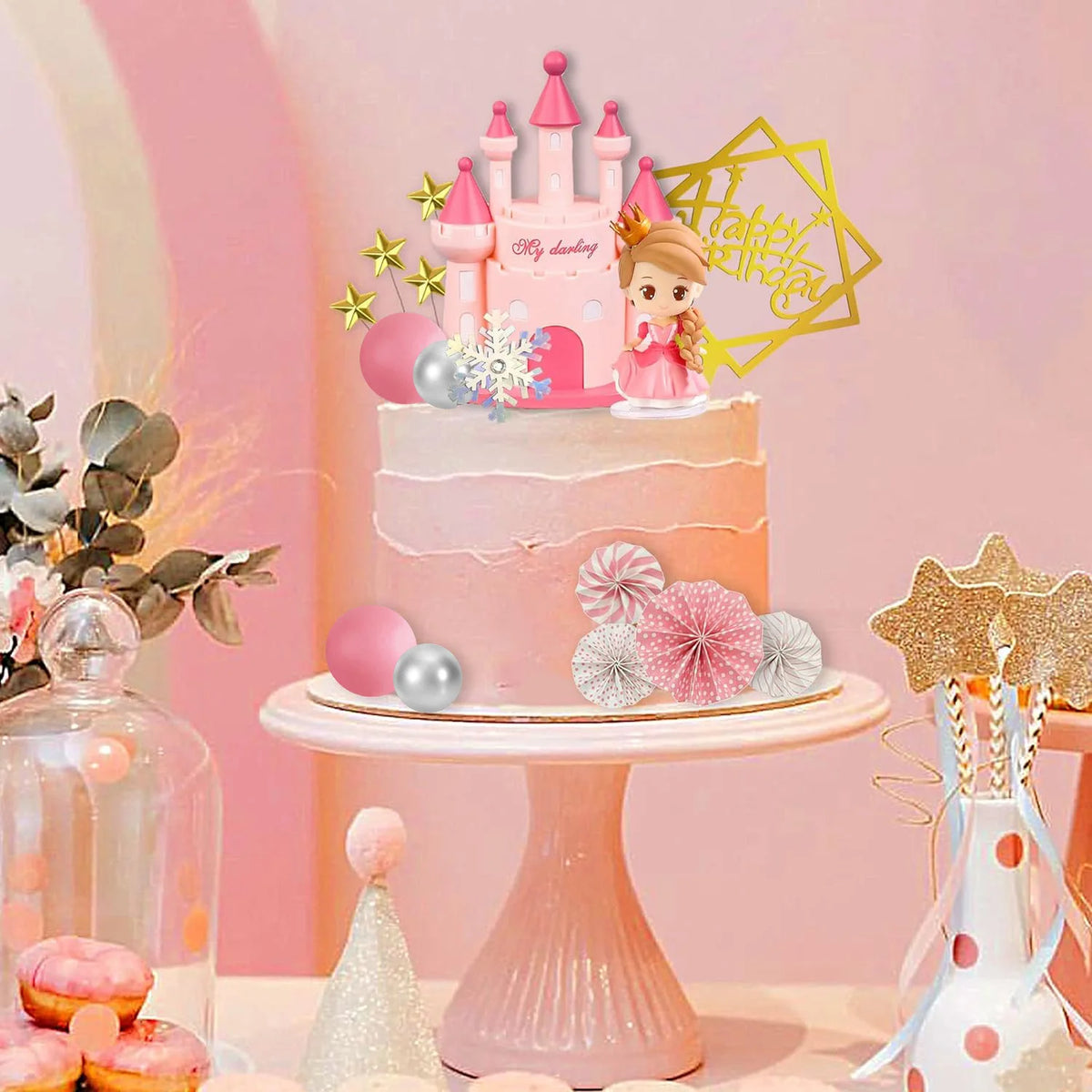 A pink tiered cake decorated with a castle topper, surrounded by floral accents and gold geometric shapes.