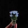 A hand holds a small glass vase filled with delicate blue glass flower sculptures.