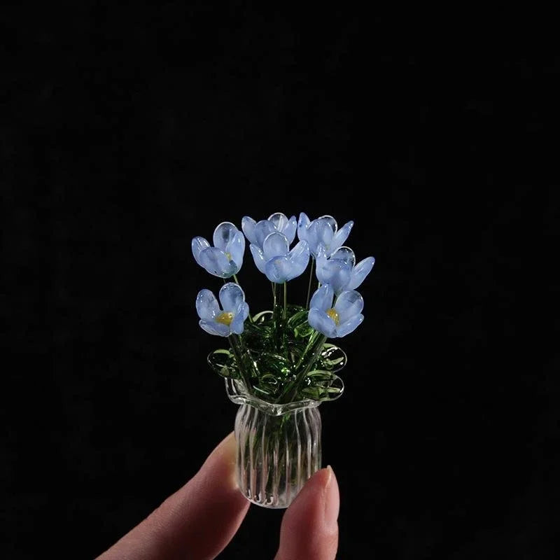 A hand holds a small glass vase filled with delicate blue glass flower sculptures.