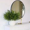 Lifelike artificial pine branches in a white vase displayed on a white mantelpiece with a round mirror.