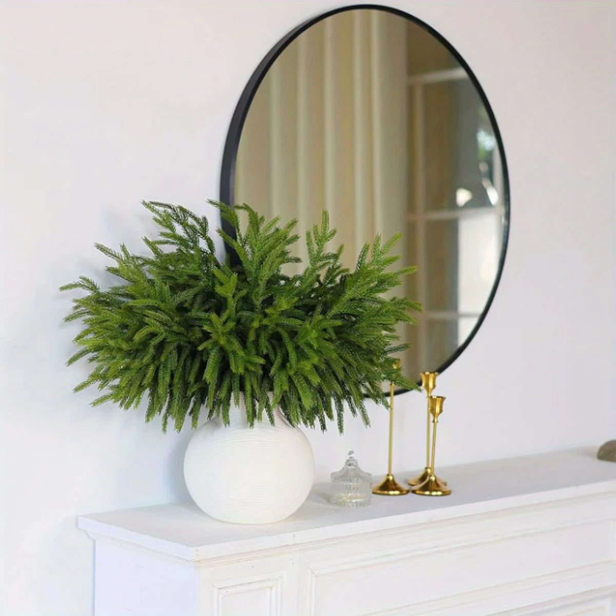 Lifelike artificial pine branches in a white vase displayed on a white mantelpiece with a round mirror.