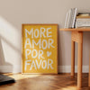 A yellow canvas wall art print with white text that reads More Amor Por Favor displayed on a wooden table.