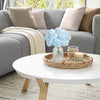 A glass vase filled with six silk blue peony flowers sits on a woven tray on a white table.