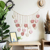 Handwoven pink leaf tassel wall hanging displayed on a light-colored wall above a wooden shelf.