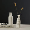 A white ceramic desktop vase with an organic ripple design holds two dried, feathery grasses against a dark background.