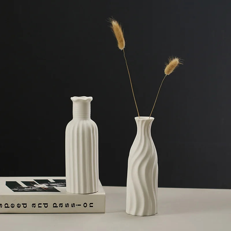 A white ceramic desktop vase with an organic ripple design holds two dried, feathery grasses against a dark background.