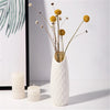 A white textured vase holds delicate dried flowers with thin stems and spherical yellow blooms.