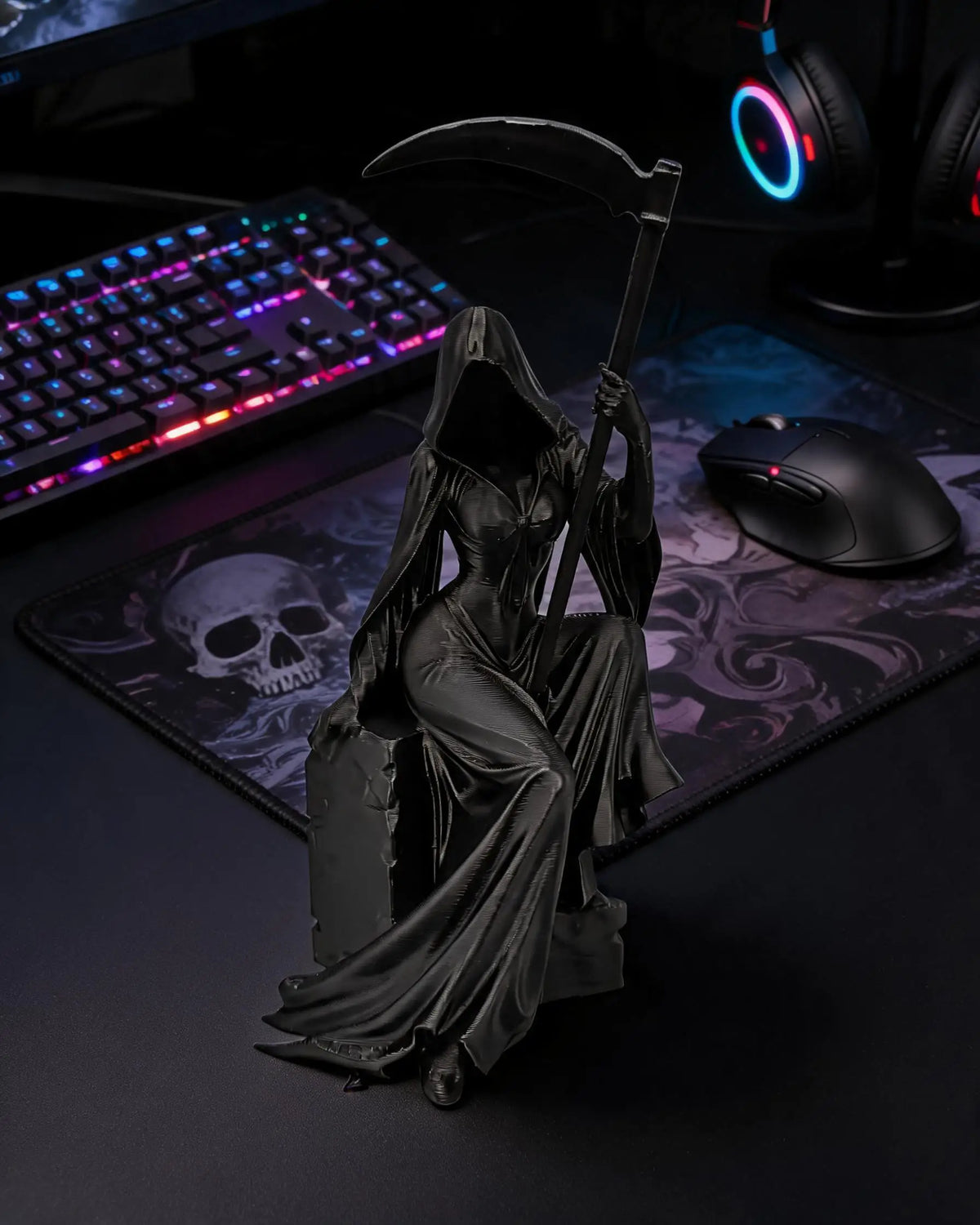 A black gothic female grim reaper figurine holding a scythe, displayed on a dark desk next to a gaming mouse and keyboard.