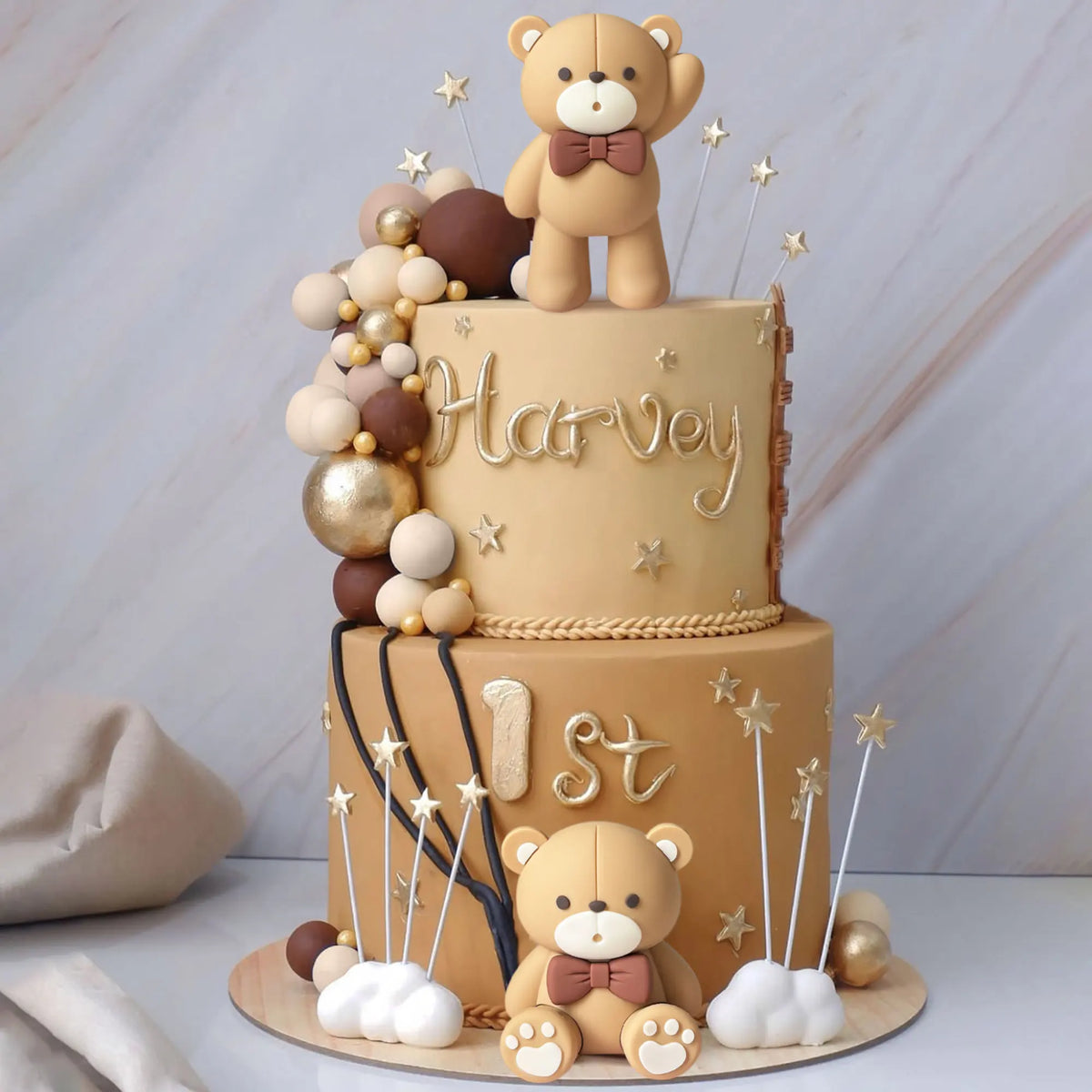 A two-tiered cake decorated with beige frosting, gold accents, and a teddy bear topper, surrounded by brown decorative balls.