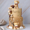 A two-tiered cake decorated with beige frosting, gold accents, and a teddy bear topper, surrounded by brown decorative balls.