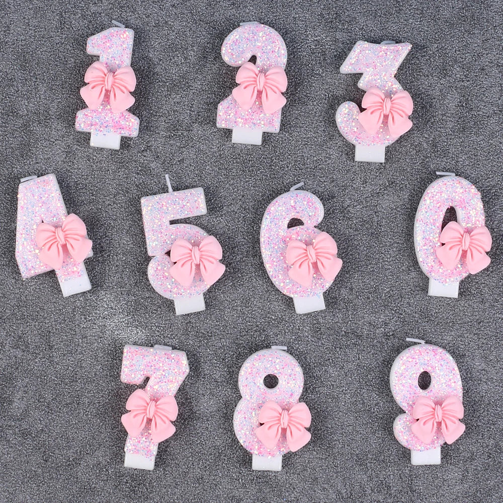 A set of glitter number cake candles from 1 to 9, each featuring a pink bow, displayed on a gray surface.