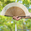 Elegant bamboo folding fan with a light beige and white pattern, held by a hand with a white tassel.