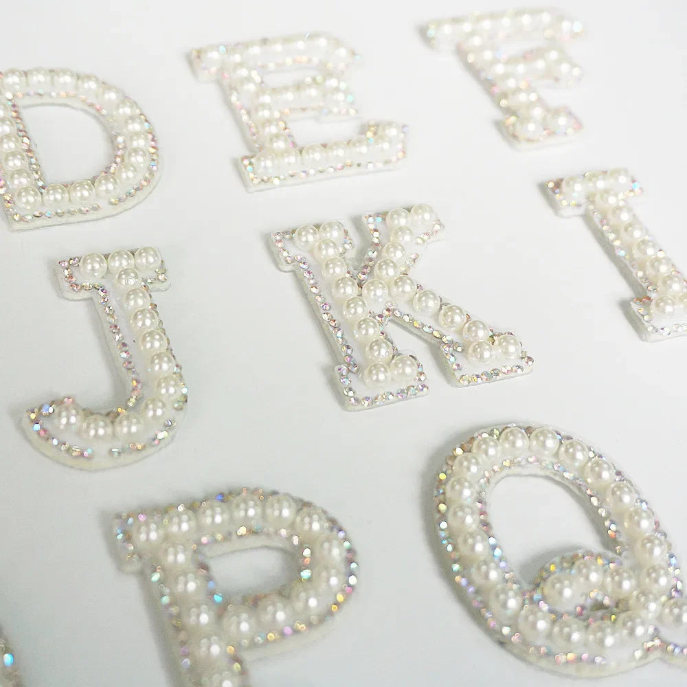 White rhinestone alphabet letters D, E, K, J, and P scattered on a white background.