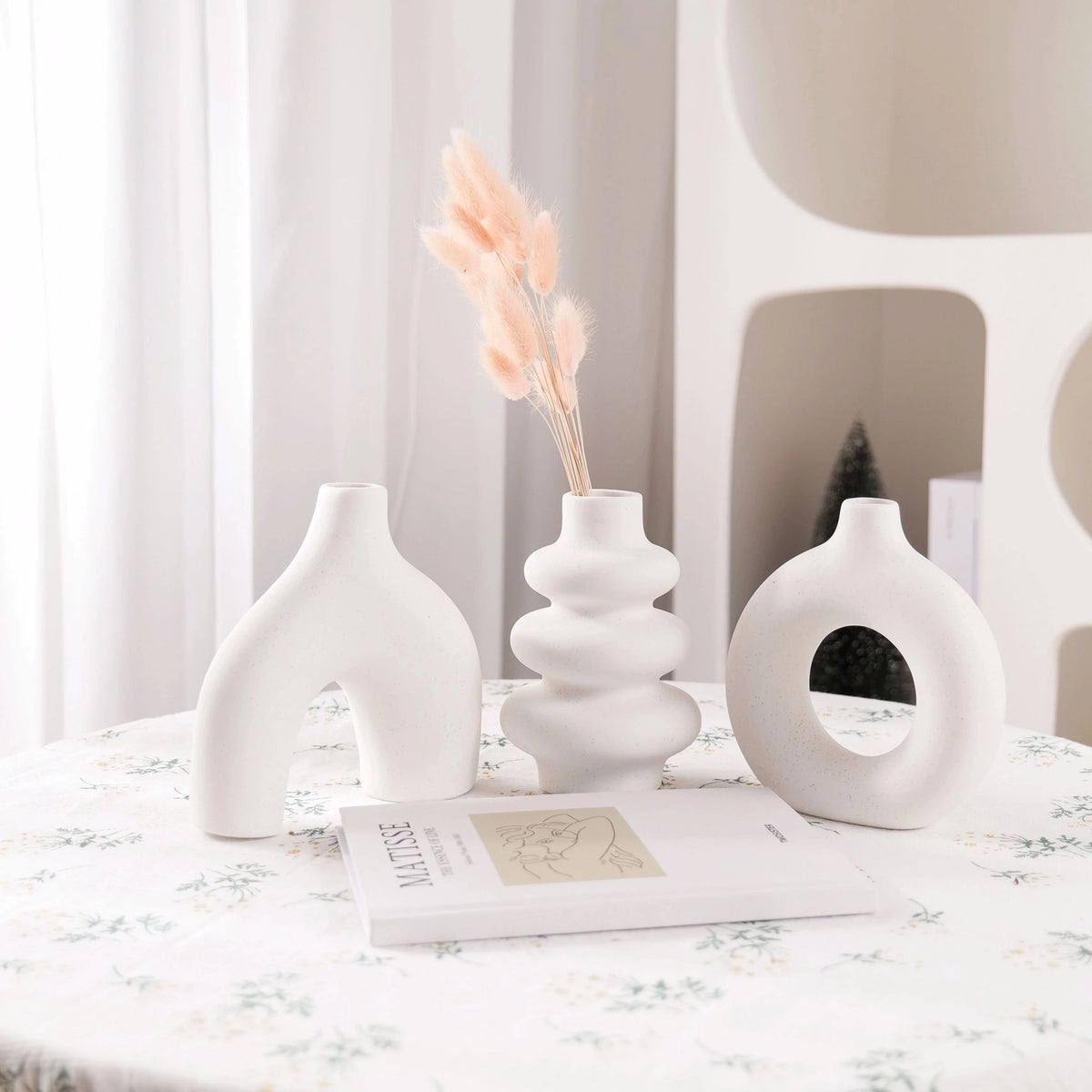 A white ceramic vase set featuring a tall floral arrangement and a circular vase displayed on a white tablecloth.