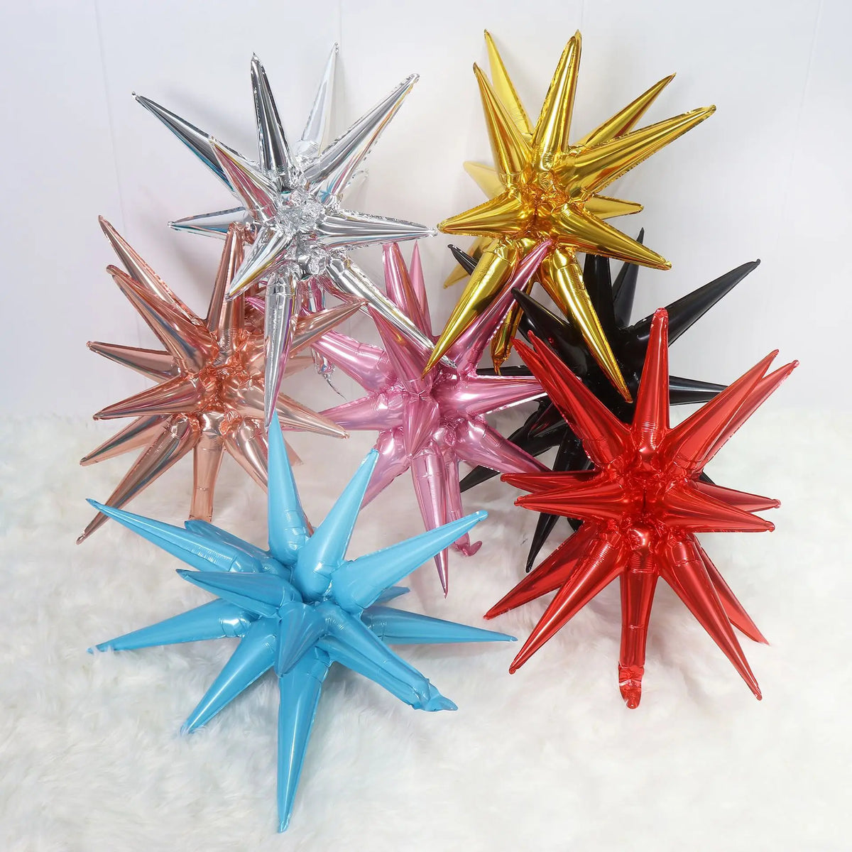 A collection of shiny foil star burst balloons in silver, gold, pink, and red are displayed on a white fluffy surface.