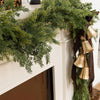 A long garland of artificial Norfolk pine greenery in winter green hangs over a white fireplace mantel.