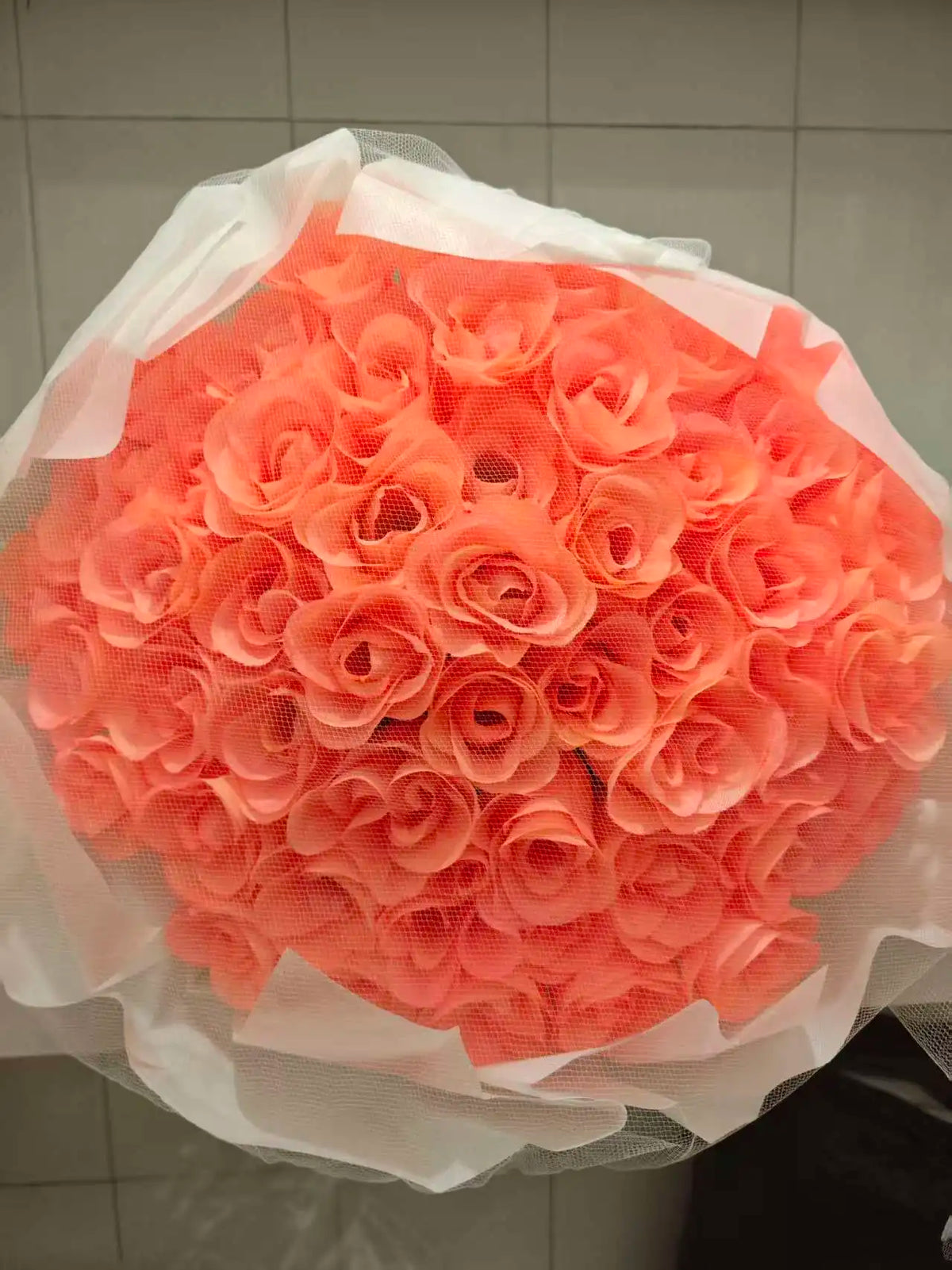 A bouquet of vibrant coral pink silk roses wrapped in sheer white tissue paper.