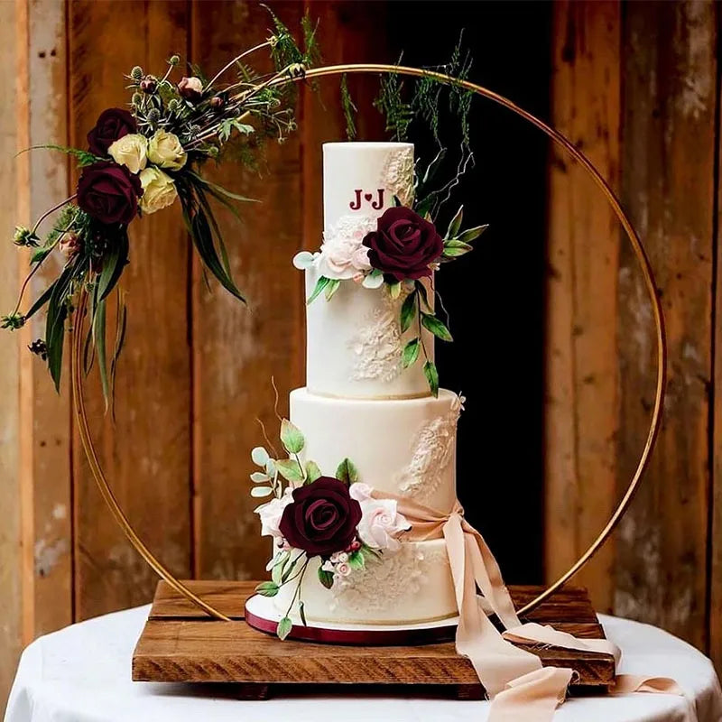 A three-tiered white cake decorated with deep red and pink roses, displayed on a wooden board with a gold hoop.