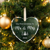 A clear heart-shaped crystal ornament with 'Our First Christmas As Mr & Mrs 2026' engraved on it, hanging on a green Christma