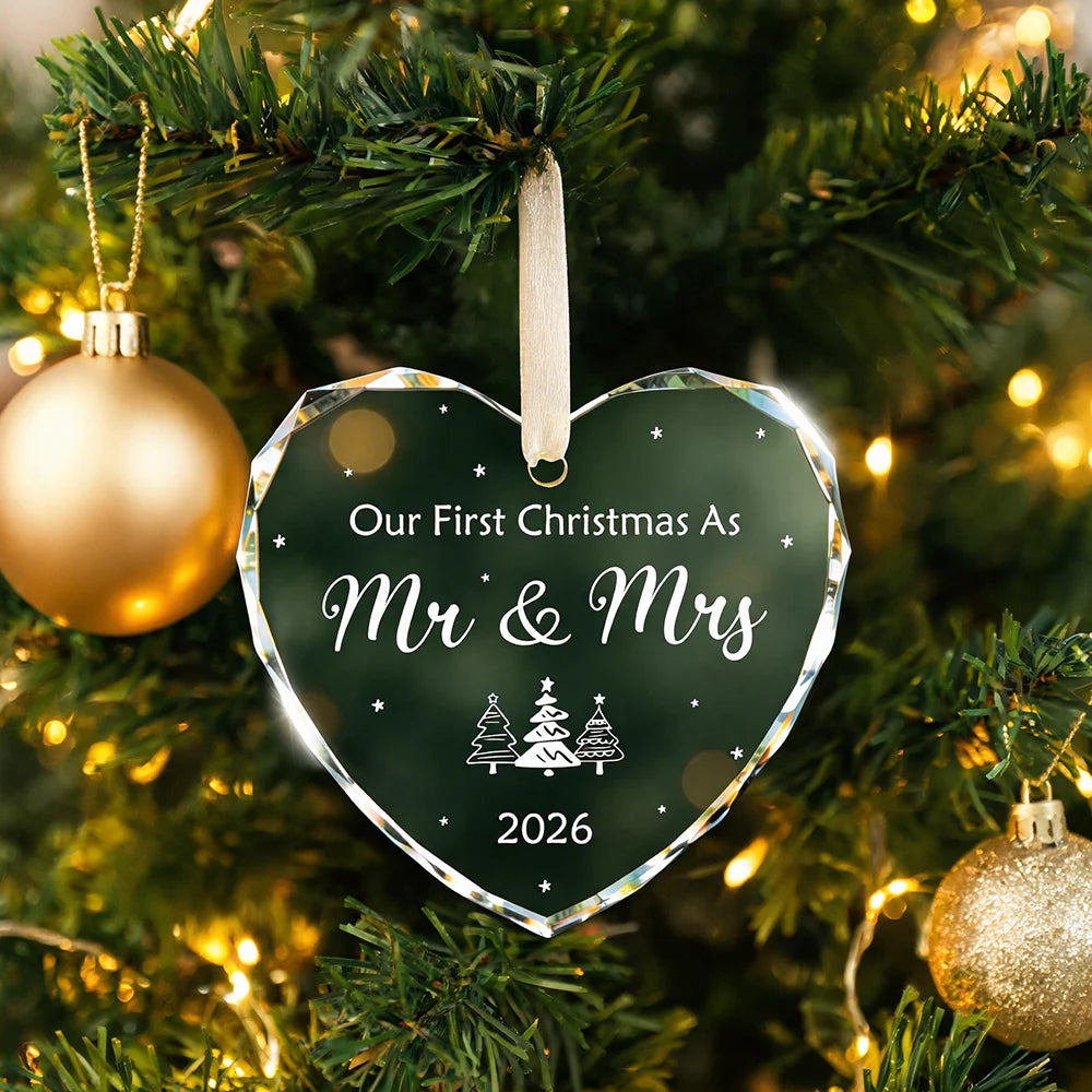 A clear heart-shaped crystal ornament with the inscription Our First Christmas As Mr & Mrs 2026 hanging on a green Christmas 