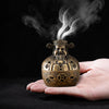 A hand holds a vintage brass incense burner with smoke rising from it against a dark background.