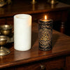 A white pillar candle and a dark metal tin candle with a flame are displayed on a wooden surface.