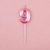 A pink number nine candle on a white stick against a light pink background.