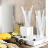 A clear glass container filled with transparent plastic drinking straws rests on a white marble surface.