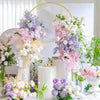 A floral arrangement display featuring pink, purple, and white flowers on a white backdrop with a gold metal arch.