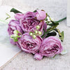 A bouquet of artificial silk roses and peonies in shades of purple and pink resting on a white surface.