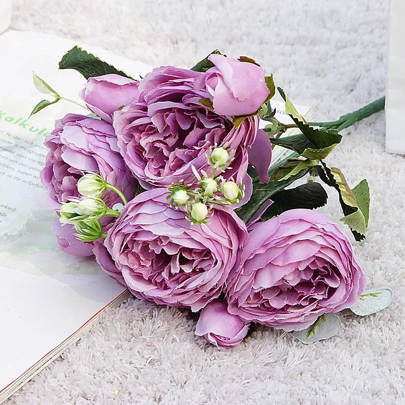 A bouquet of artificial silk roses and peonies in shades of purple and pink resting on a white surface.