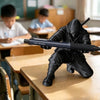 A black ninja figure is posed on a wooden desk, holding a black pen, in a classroom setting.
